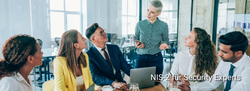 Personengruppe in Besprechungssituation mit Laptop und Getränken, eine Person präsentiert stehend, eingeblendeter Text „NIS2-Praxis Workshop“ – Darstellung eines interaktiven Workshops zur praktischen Umsetzung der NIS2-Richtlinie.