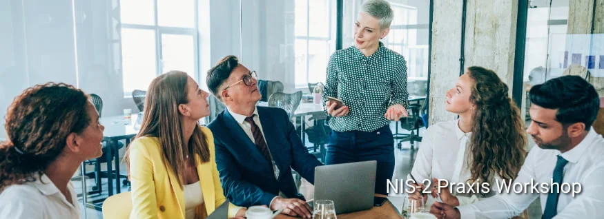 Personengruppe in Besprechungssituation mit Laptop und Getränken, eine Person präsentiert stehend, eingeblendeter Text „NIS2-Praxis Workshop“ – Darstellung eines interaktiven Workshops zur praktischen Umsetzung der NIS2-Richtlinie.