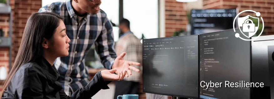 Zwei Personen vor Monitoren mit Codeanzeigen in Büroumgebung, ergänzt durch Schloss-Symbol – Darstellung für Cyber Resilience oder IT-Sicherheitsmaßnahmen im Arbeitsalltag.